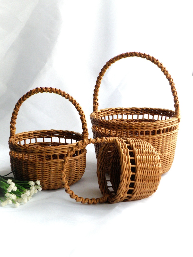 May include: Three brown woven baskets of different sizes, each with an arched handle. The baskets are made from a light brown material and feature a woven pattern. The handles are also woven and curved. They are arranged on a white surface.