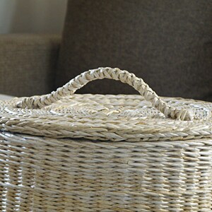 May include: A white wicker basket with a woven lid and a handle. The basket is made of natural materials and has a rustic look.