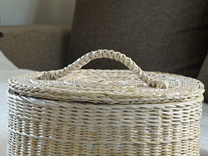 May include: A white wicker basket with a woven lid and a handle. The basket is made of natural materials and has a rustic look.