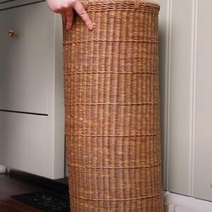 May include: A tall, cylindrical umbrella stand crafted from light brown woven wicker. The stand features a consistent, tight weave pattern throughout its vertical structure, providing a sturdy and open-top design for holding umbrellas or walking sticks.