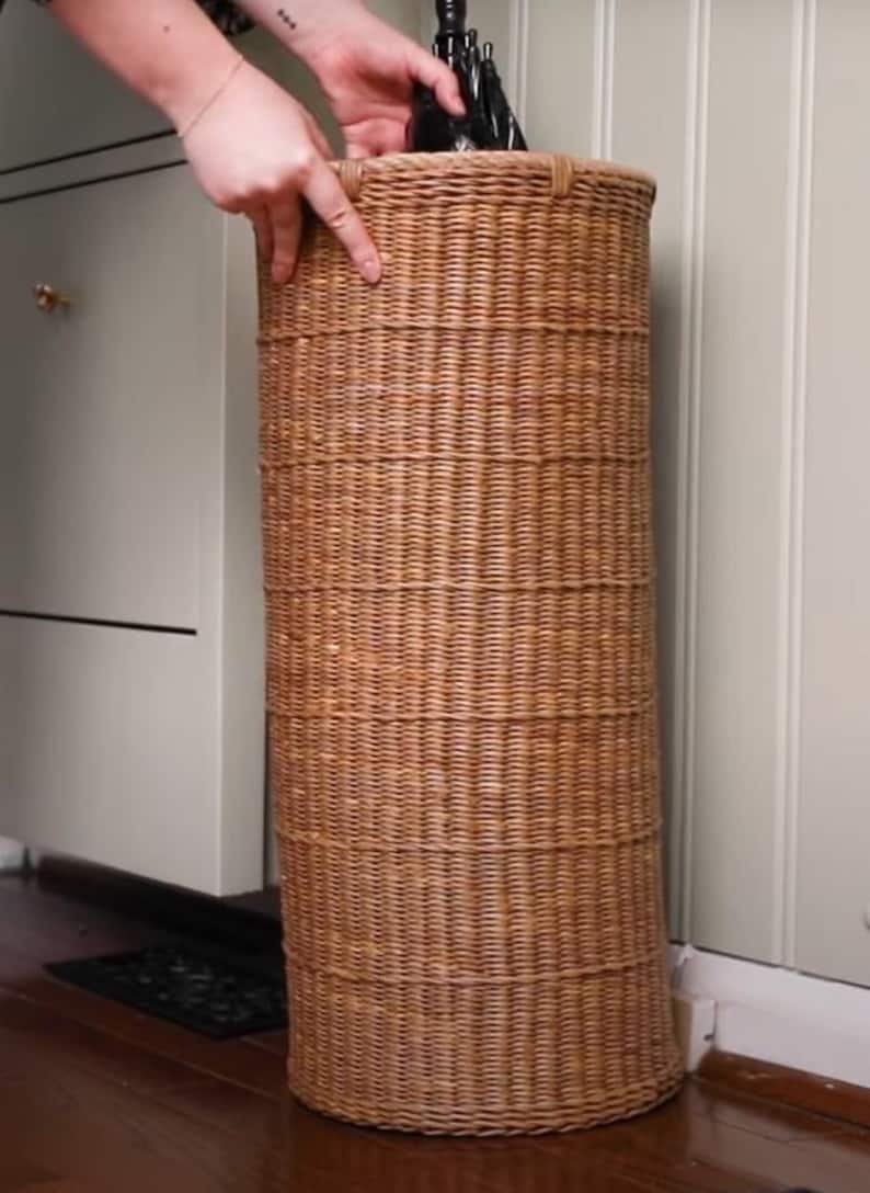 May include: A tall, cylindrical umbrella stand crafted from light brown woven wicker. The stand features a consistent, tight weave pattern throughout its vertical structure, providing a sturdy and open-top design for holding umbrellas or walking sticks.