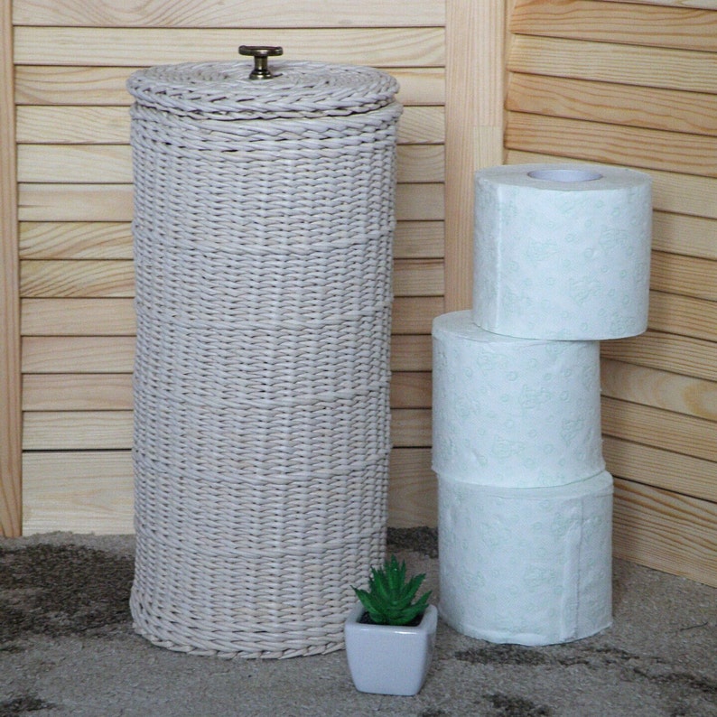 May include: White wicker toilet paper holder with a brass knob on top. Three rolls of white toilet paper are stacked next to the holder. A small potted succulent plant is in front of the holder.