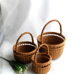 May include: Three brown woven baskets of different sizes, each with a rounded handle. The baskets are displayed together against a white backdrop. The smallest basket is in the foreground, with the largest basket behind.