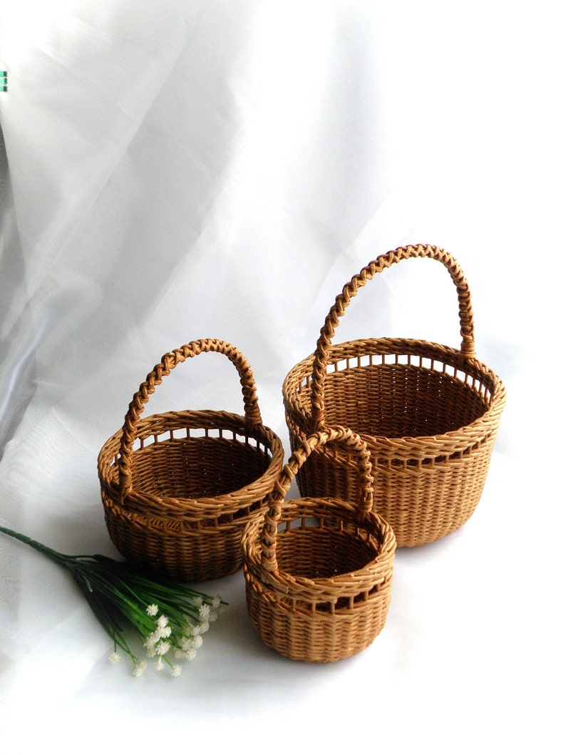 May include: Three brown woven baskets of different sizes, each with a rounded handle. The baskets are displayed together against a white backdrop. The smallest basket is in the foreground, with the largest basket behind.