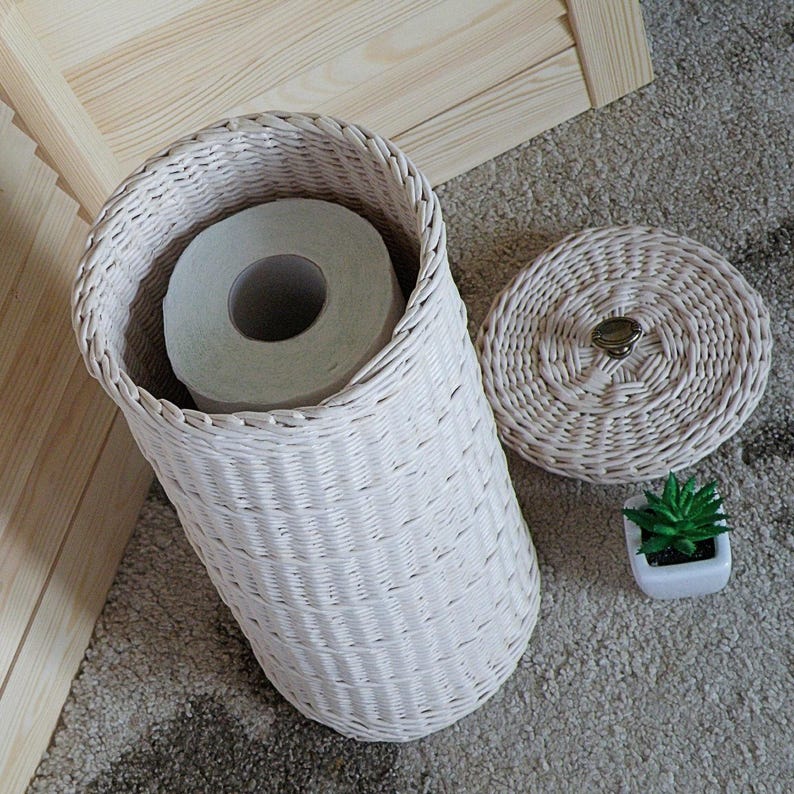 May include: White wicker toilet paper holder with a roll of toilet paper inside and a matching lid. The holder is sitting on a beige carpet.