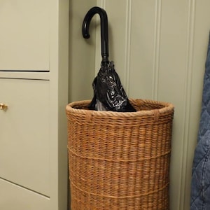 May include: A tall, cylindrical woven wicker basket with a natural brown finish and a thick braided rim. Inside the basket stands a black umbrella with a curved handle, its fabric canopy folded and tucked neatly within the woven structure.