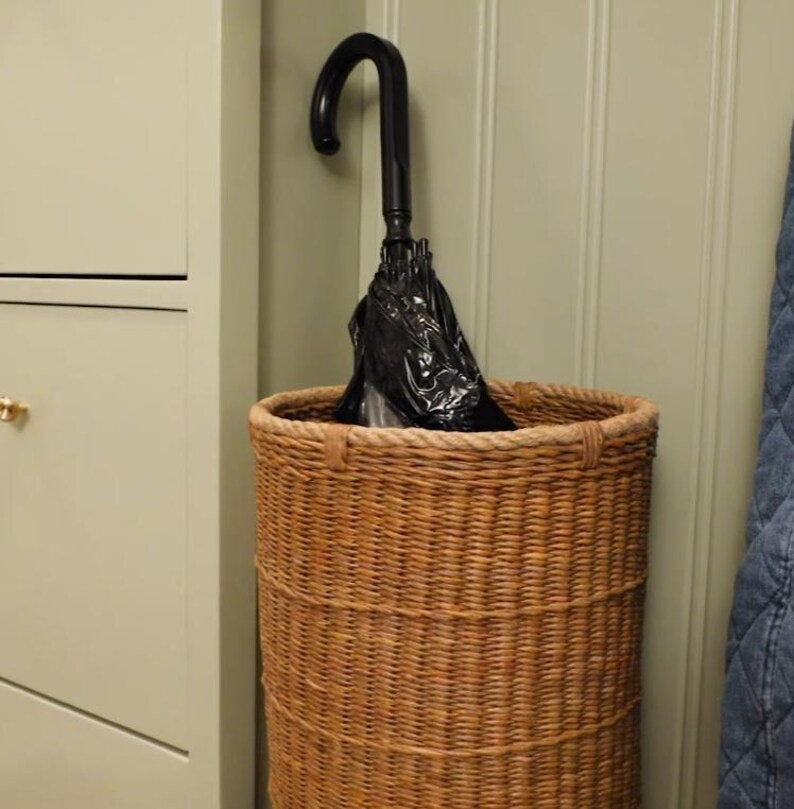 May include: A tall, cylindrical woven wicker basket with a natural brown finish and a thick braided rim. Inside the basket stands a black umbrella with a curved handle, its fabric canopy folded and tucked neatly within the woven structure.