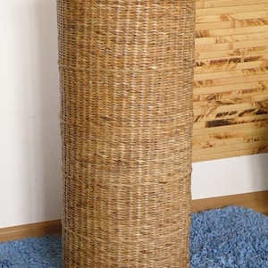 May include: A tall, cylindrical basket woven from light brown natural fibres. The structure features a tight, vertical weave pattern throughout the body, with a thicker, braided rope-like rim encircling the top edge of the open container.