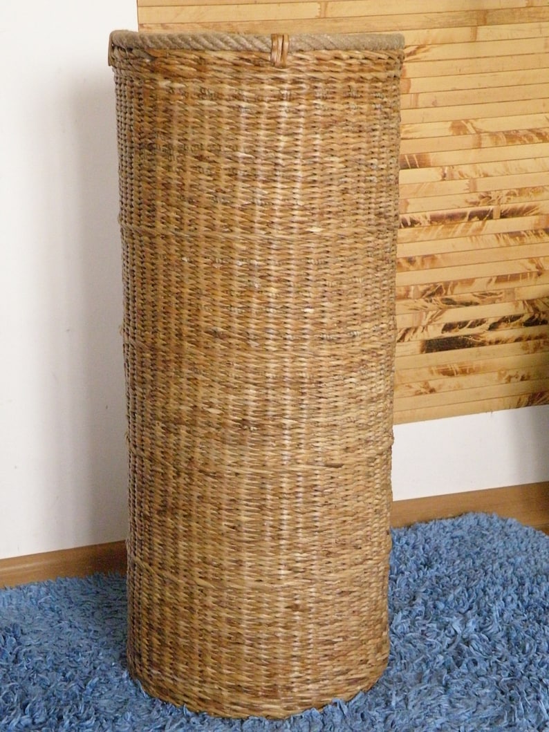 May include: A tall, cylindrical basket woven from light brown natural fibres. The structure features a tight, vertical weave pattern throughout the body, with a thicker, braided rope-like rim encircling the top edge of the open container.