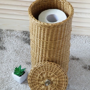 Puede incluir: Un porta rollo de papel higi&eacute;nico de mimbre tejido con un rollo de papel higi&eacute;nico dentro y una tapa con un pomo de metal. El porta rollo est&aacute; sobre una alfombra beige.