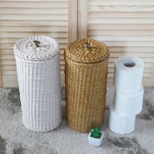 Puede incluir: Dos porta rollos de papel higi&eacute;nico de mimbre tejido, uno blanco y otro marr&oacute;n, con una peque&ntilde;a planta en maceta en frente. A la derecha hay una pila de tres rollos de papel higi&eacute;nico blancos.