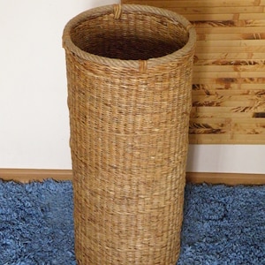 May include: A tall, cylindrical woven basket made of natural light brown fibres. The basket features a sturdy, thick rope trim around the circular top opening, providing a textured finish to the simple, vertical design of the woven structure.