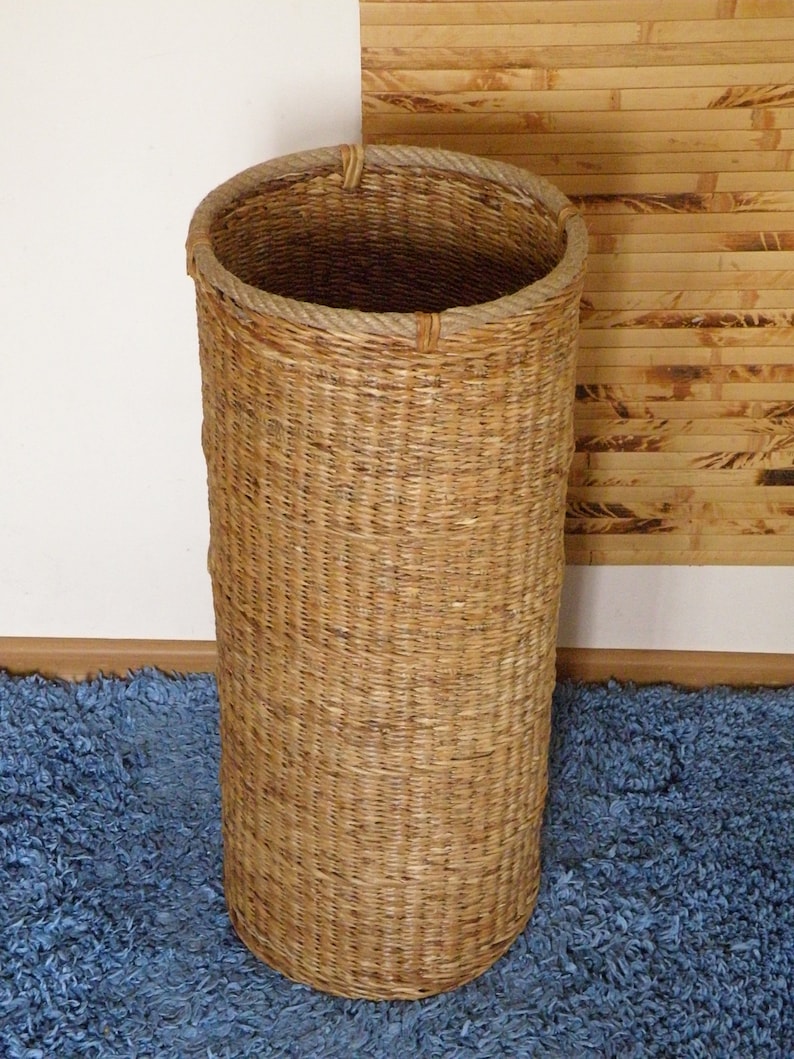 May include: A tall, cylindrical basket woven from natural light brown fibres. The basket features a vertical weave pattern and a thick, braided rope trim encircling the top rim, providing a sturdy edge to the open-topped, hollow structure.