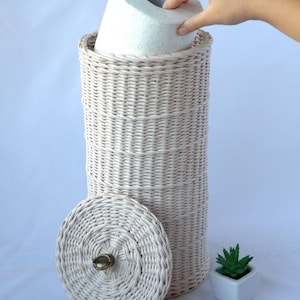 Puede incluir: Porta rollo de papel higi&eacute;nico de mimbre blanco con tapa y un rollo de papel higi&eacute;nico dentro. El porta rollo tiene un pomo de lat&oacute;n en la tapa.