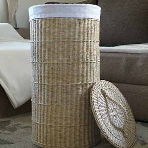 May include: A woven wicker laundry basket with a white fabric liner and a matching lid. The basket is sitting on a patterned rug.