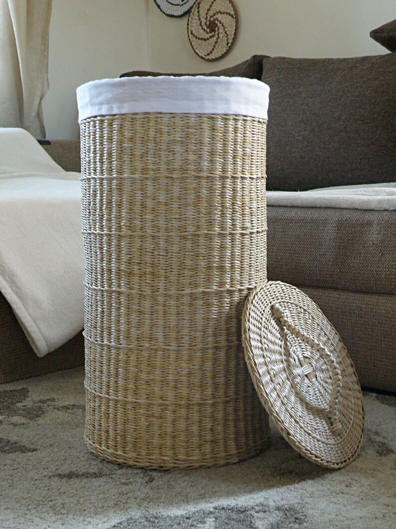 May include: A woven wicker laundry basket with a white fabric liner and a matching lid. The basket is sitting on a patterned rug.