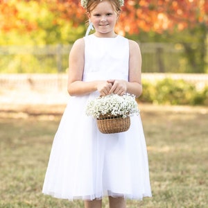 May include: A person in a white sleeveless dress and a flower crown, carrying a woven basket of white flowers. The background shows a blurred tree with red and orange leaves, typical of autumn.