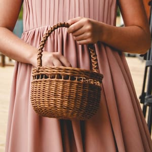 May include: A small, woven brown wicker basket with a braided handle. The basket is held by a person wearing a pink dress. The basket is likely used for carrying flowers or small items.