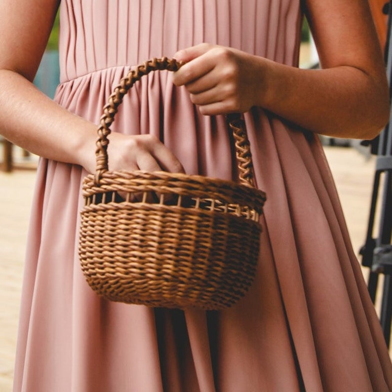 May include: A small, woven brown wicker basket with a braided handle. The basket is held by a person wearing a pink dress. The basket is likely used for carrying flowers or small items.