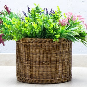 May include: A brown wicker basket filled with a variety of green and purple flowers. The basket is round and has a woven texture.