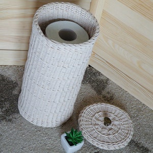 May include: White wicker toilet paper holder with a roll of toilet paper inside and a matching lid. The holder is sitting on a brown carpet.