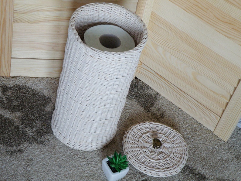 May include: White wicker toilet paper holder with a roll of toilet paper inside and a matching lid. The holder is sitting on a brown carpet.