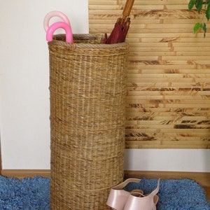 May include: A tall, cylindrical woven wicker umbrella stand with a thick rope rim. Inside are two small pink umbrellas and one large umbrella with a dark red canopy and a curved wooden handle. A pair of light pink heeled sandals sits in front.