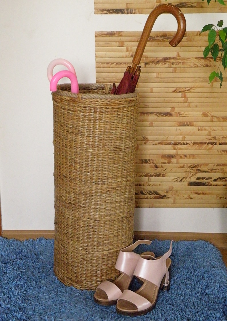 May include: A tall, cylindrical woven wicker umbrella stand with a thick rope rim. Inside are two small pink umbrellas and one large umbrella with a dark red canopy and a curved wooden handle. A pair of light pink heeled sandals sits in front.