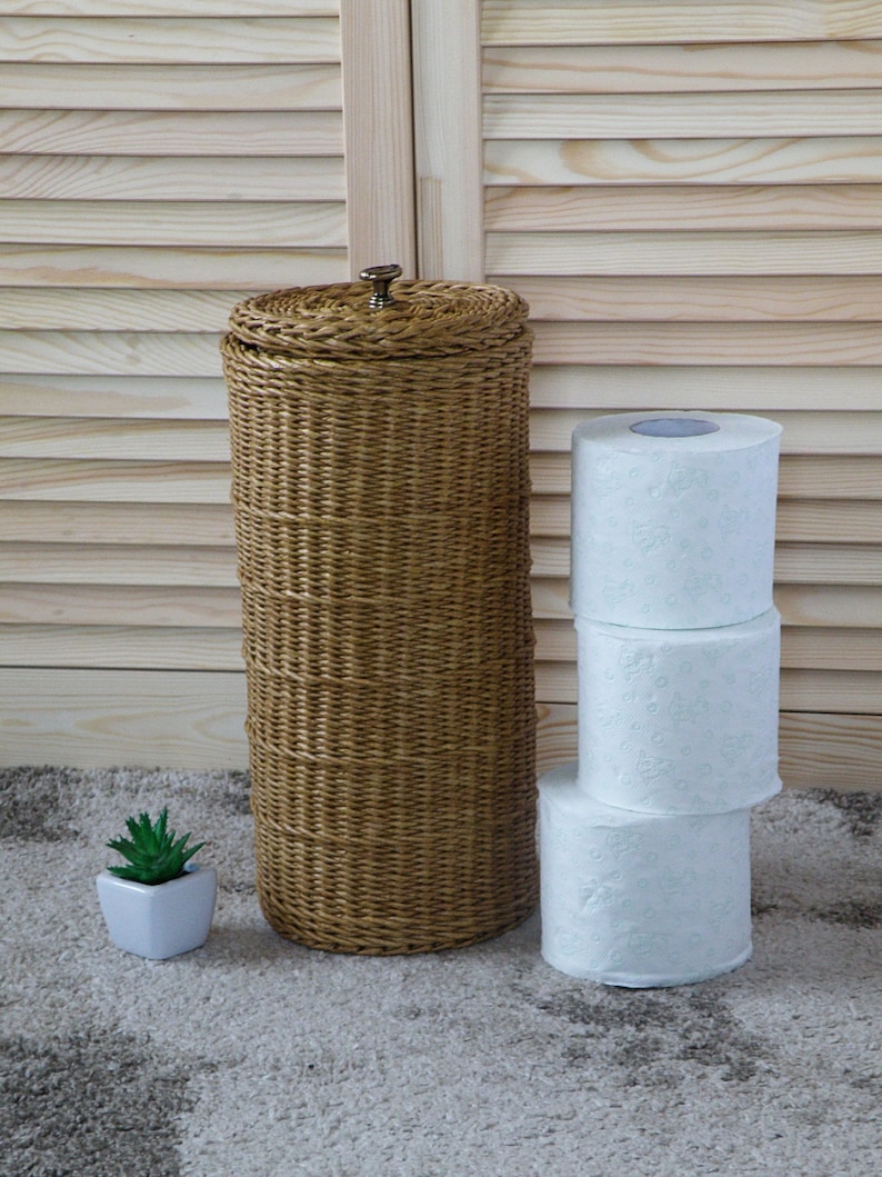 May include: A wicker toilet paper holder with a lid and a stack of three white toilet paper rolls. A small potted succulent plant is in the foreground.