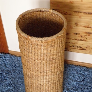 May include: A tall, cylindrical woven basket with a light brown natural finish. The basket features a thick, braided rope trim around the circular top opening, providing a sturdy edge to the woven structure of the tall, hollow container.