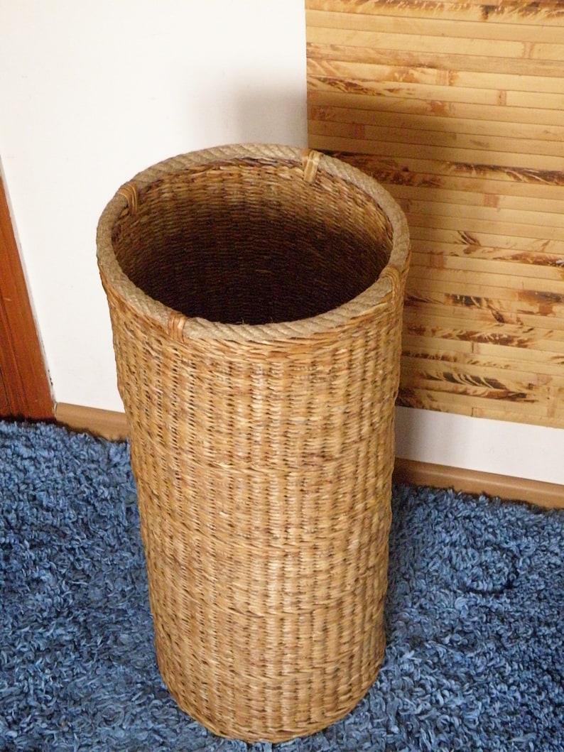 May include: A tall, cylindrical woven basket with a light brown natural finish. The basket features a thick, braided rope trim around the circular top opening, providing a sturdy edge to the woven structure of the tall, hollow container.