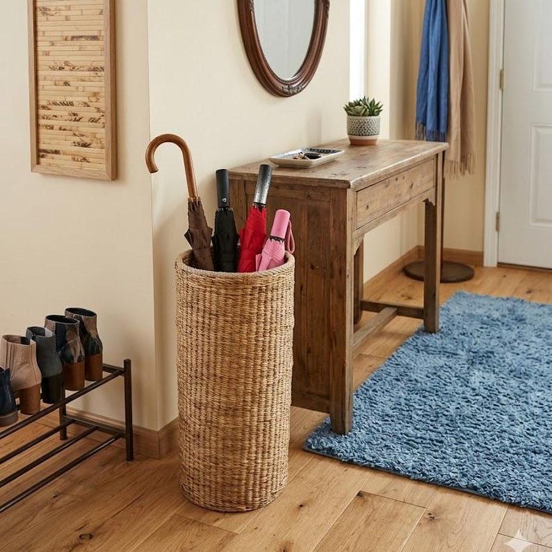 May include: A cylindrical woven wicker umbrella stand with a natural tan finish. The stand holds four umbrellas, including one with a curved wooden handle, one black umbrella, one red umbrella, and one light pink umbrella, all standing upright.