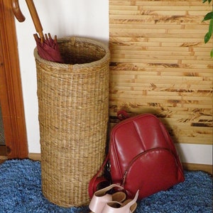 May include: A tall cylindrical woven wicker umbrella stand containing a burgundy umbrella with a curved wooden handle, a red leather backpack with a front zippered pocket, and a pair of light pink open-toe sandals with block heels.