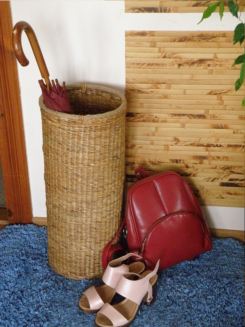 May include: A tall cylindrical woven wicker umbrella stand containing a burgundy umbrella with a curved wooden handle, a red leather backpack with a front zippered pocket, and a pair of light pink open-toe sandals with block heels.