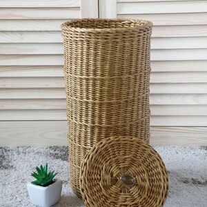 May include: A woven wicker storage basket with a lid. The basket is brown and has a natural finish. The lid has a circular design and a metal handle.