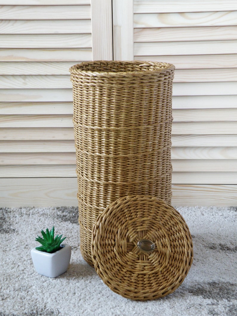 May include: A woven wicker storage basket with a lid. The basket is brown and has a natural finish. The lid has a circular design and a metal handle.