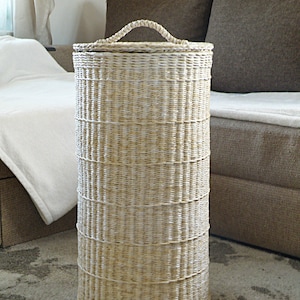 May include: A large, woven wicker basket with a lid and a handle. The basket is light brown and has a natural, rustic look.
