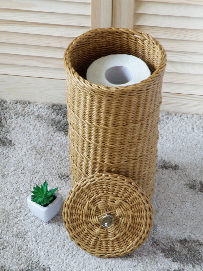 May include: A woven wicker toilet paper holder with a roll of toilet paper inside and a matching lid. The holder has a decorative knob on the lid. A small potted succulent plant is in the foreground.