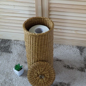 May include: A woven wicker toilet paper holder with a lid and a roll of toilet paper inside. The holder is sitting on a light brown rug.