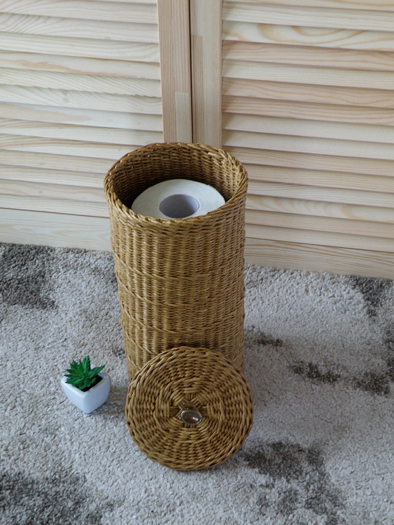 May include: A woven wicker toilet paper holder with a lid and a roll of toilet paper inside. The holder is sitting on a light brown rug.
