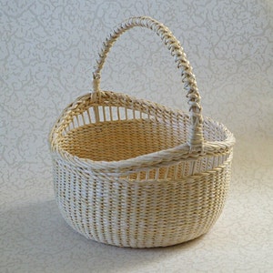 May include: A white wicker basket with a woven handle. The basket is oval shaped and has a wide opening.