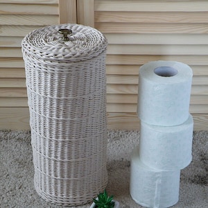 May include: White wicker toilet paper holder with a lid and a brass knob. The holder is filled with three rolls of white toilet paper.
