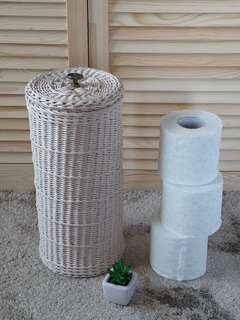 May include: White wicker toilet paper holder with a lid and a brass knob. The holder is filled with three rolls of white toilet paper.
