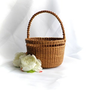 May include: A small, round, woven basket with a braided handle. The basket is light brown and sits next to two white artificial flowers with green leaves. The background is white.