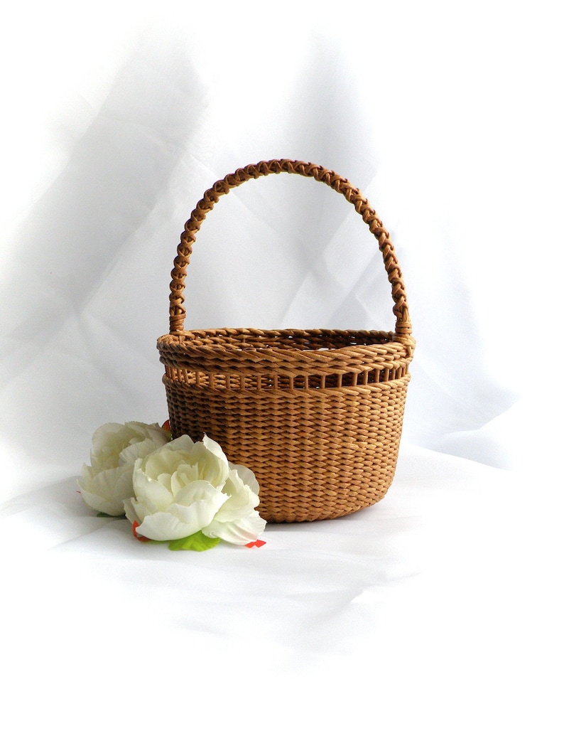 May include: A small, round, woven basket with a braided handle. The basket is light brown and sits next to two white artificial flowers with green leaves. The background is white.