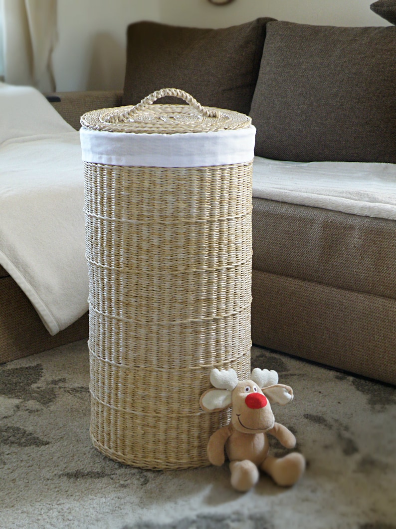 May include: A woven wicker laundry basket with a white fabric lid. The basket is standing on a light brown rug with a plush brown reindeer toy sitting in front of it.