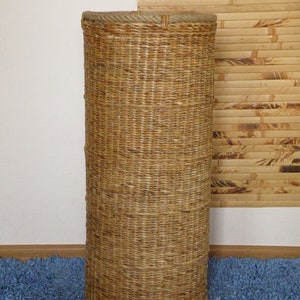 May include: A tall, cylindrical basket woven from natural light brown fibres. The structure features a tight, vertical weave pattern with a reinforced, thicker braided rim at the top edge, creating a sturdy, upright container with a consistent diameter.