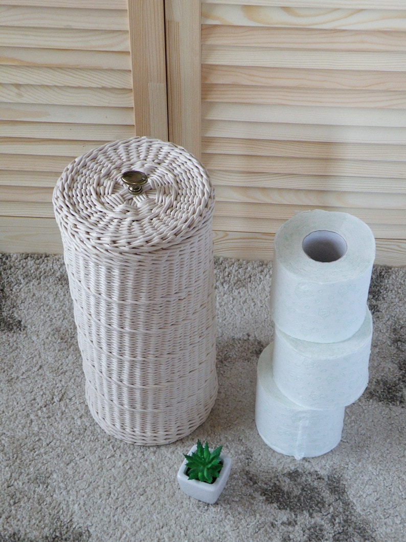 May include: A white wicker toilet paper holder with a brass knob on top. Three rolls of white toilet paper are stacked next to the holder. A small potted succulent plant is in the foreground.