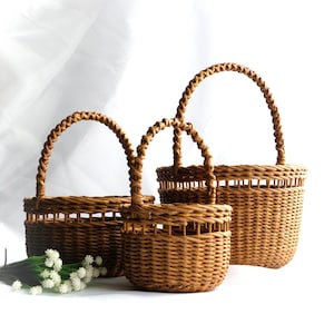 May include: Three woven wicker baskets in different sizes, featuring arched handles. The baskets are a warm brown hue and have a textured finish. Small white flowers are in front of the baskets.