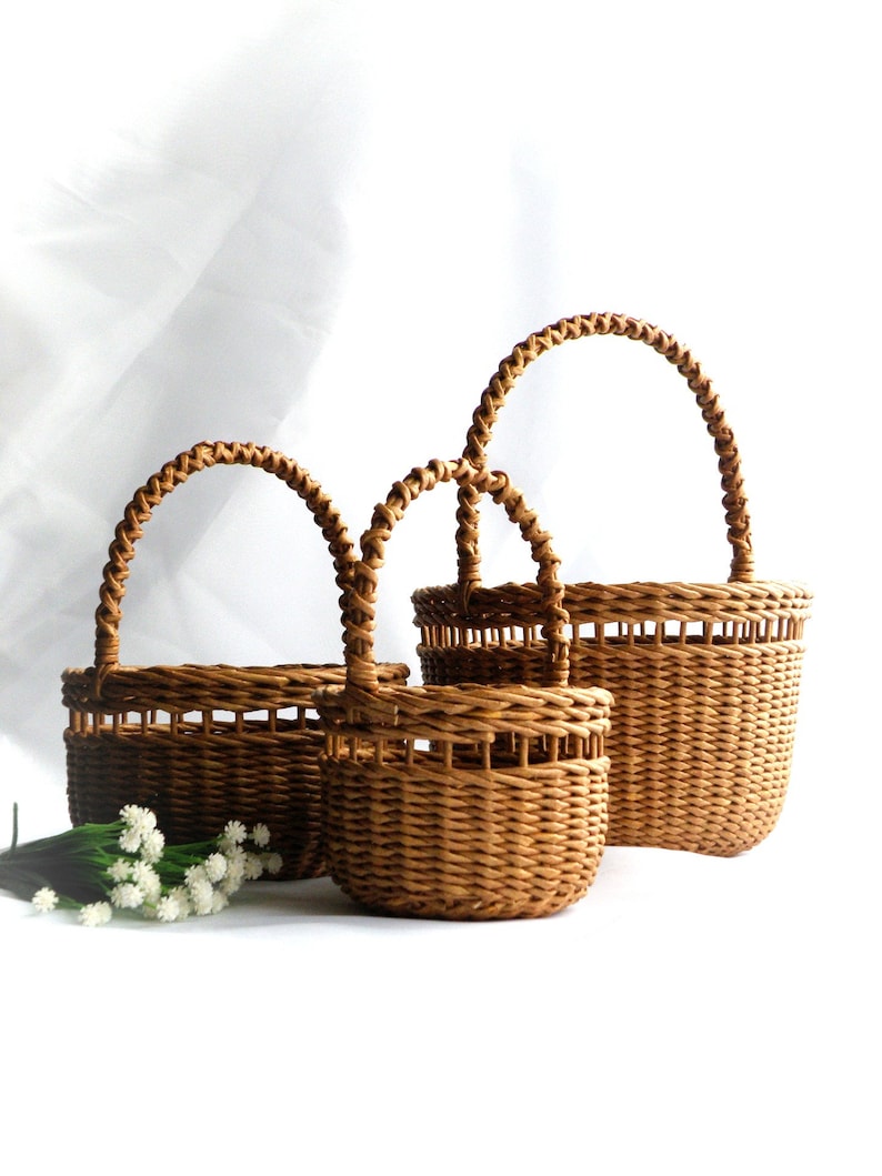 May include: Three woven wicker baskets in different sizes, featuring arched handles. The baskets are a warm brown hue and have a textured finish. Small white flowers are in front of the baskets.